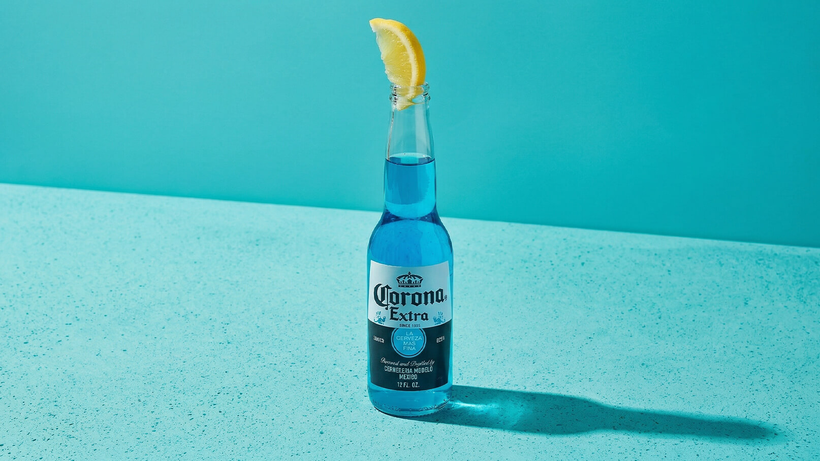 Eine Flasche Corona Extra steht auf einem blauen Untergrund. Die Flasche ist teilweise mit blauer Flüssigkeit gefüllt und hat eine Zitronenscheibe auf dem Deckel.