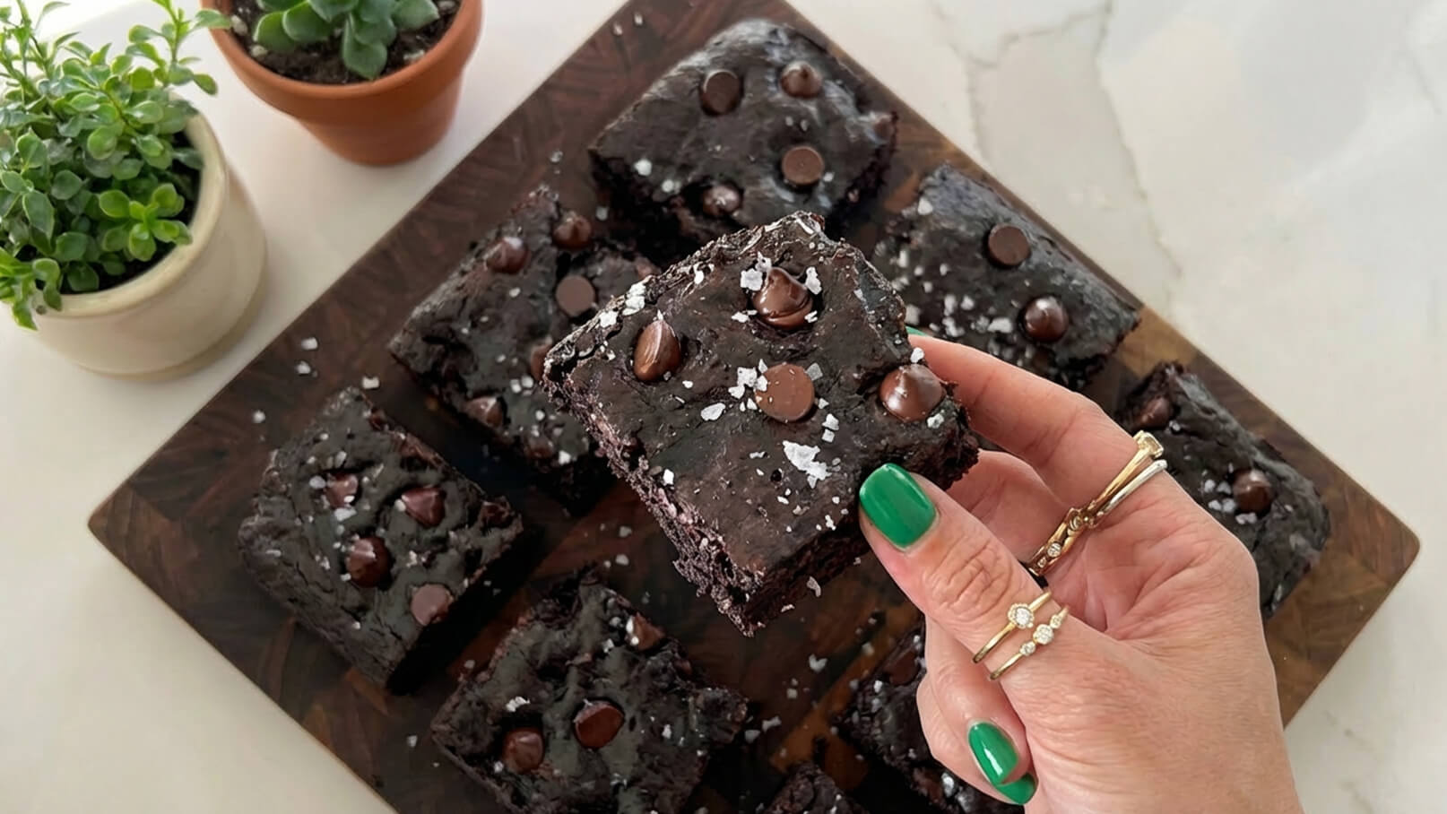 Eine Hand hält ein Stück schokoladigen Brownies mit Schokoladenstückchen und einer Prise Salz. Im Hintergrund sind mehrere Brownies auf einem Holzbrett angeordnet. Grünes Nagellack ist sichtbar, ebenso wie einige kleine Topfpflanzen.