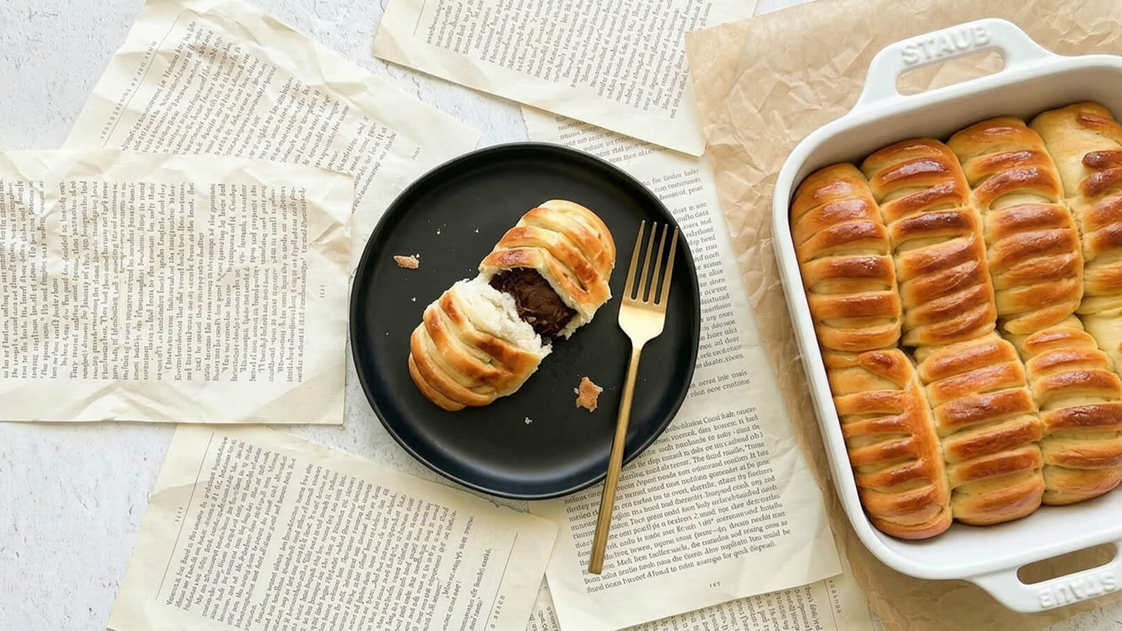 Ein schwarzes Teller mit einem aufgeplatzten, gestrichenen Gebäckstück und einer goldenen Gabel daneben. Im Hintergrund befindet sich eine Auflaufform mit mehreren weiteren, ähnlichen Gebäckstücken. Das gesamte Arrangement ist auf einer Unterlage aus alten Buchseiten platziert.