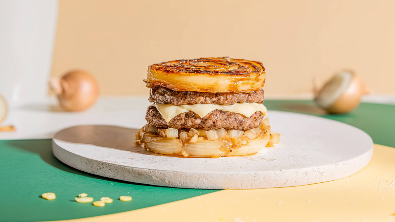 Ein gefüllter Burger aus zwei Rindfleisch-Patties und Käse, geschichtet mit gebratenen Zwiebeln. Die Zwiebeln sind auf der Ober- und Unterseite des Burgers angeordnet. Der Burger steht auf einem runden, hellen Teller vor einem farbigen Hintergrund.
