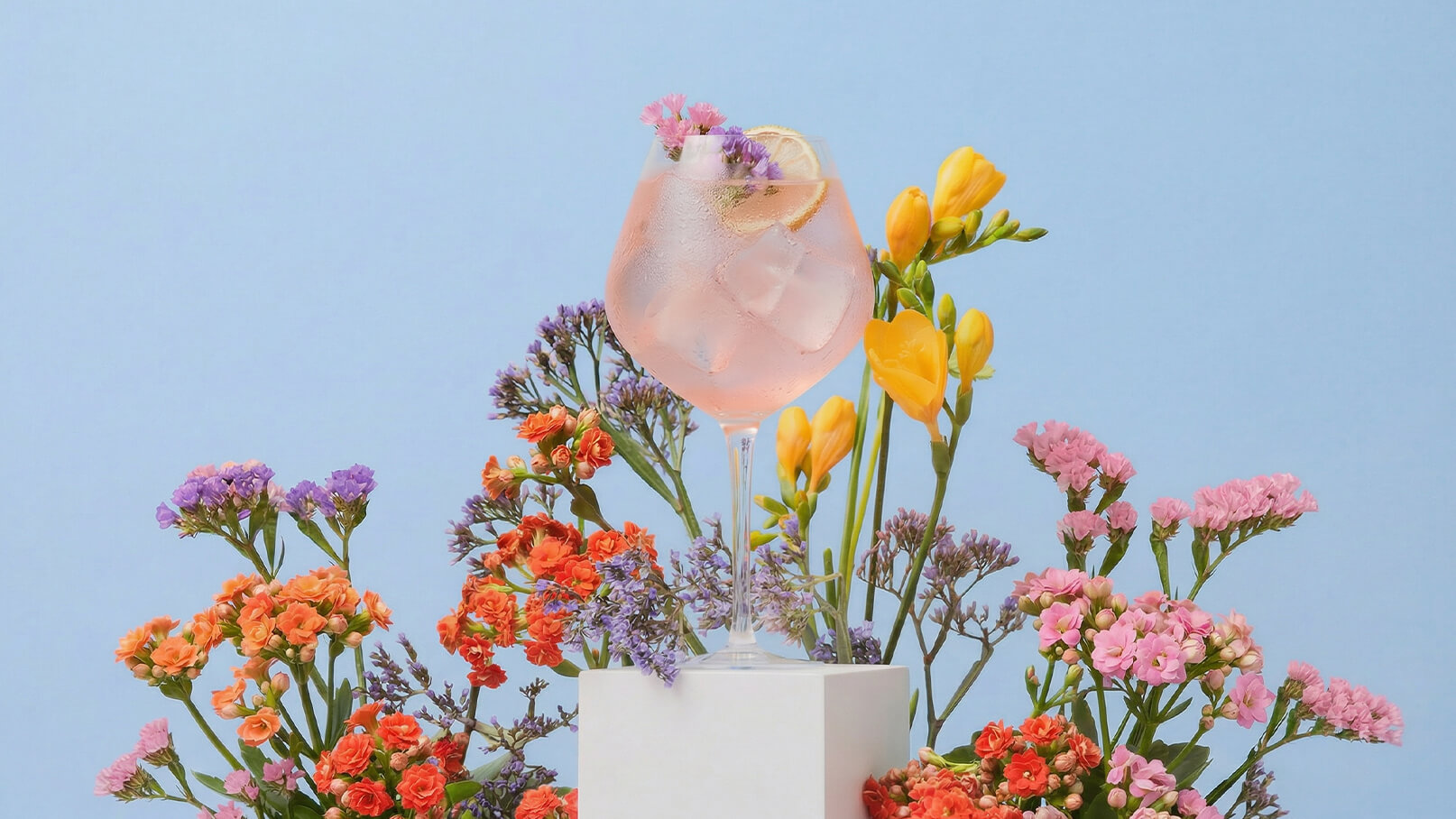 Ein elegantes Glas mit einem hellrosa Getränk steht auf einer weißen Plattform, umgeben von bunten Blumen in verschiedenen Farben. Der Hintergrund ist in einem sanften Blau gehalten, was eine fröhliche und einladende Atmosphäre schafft.
