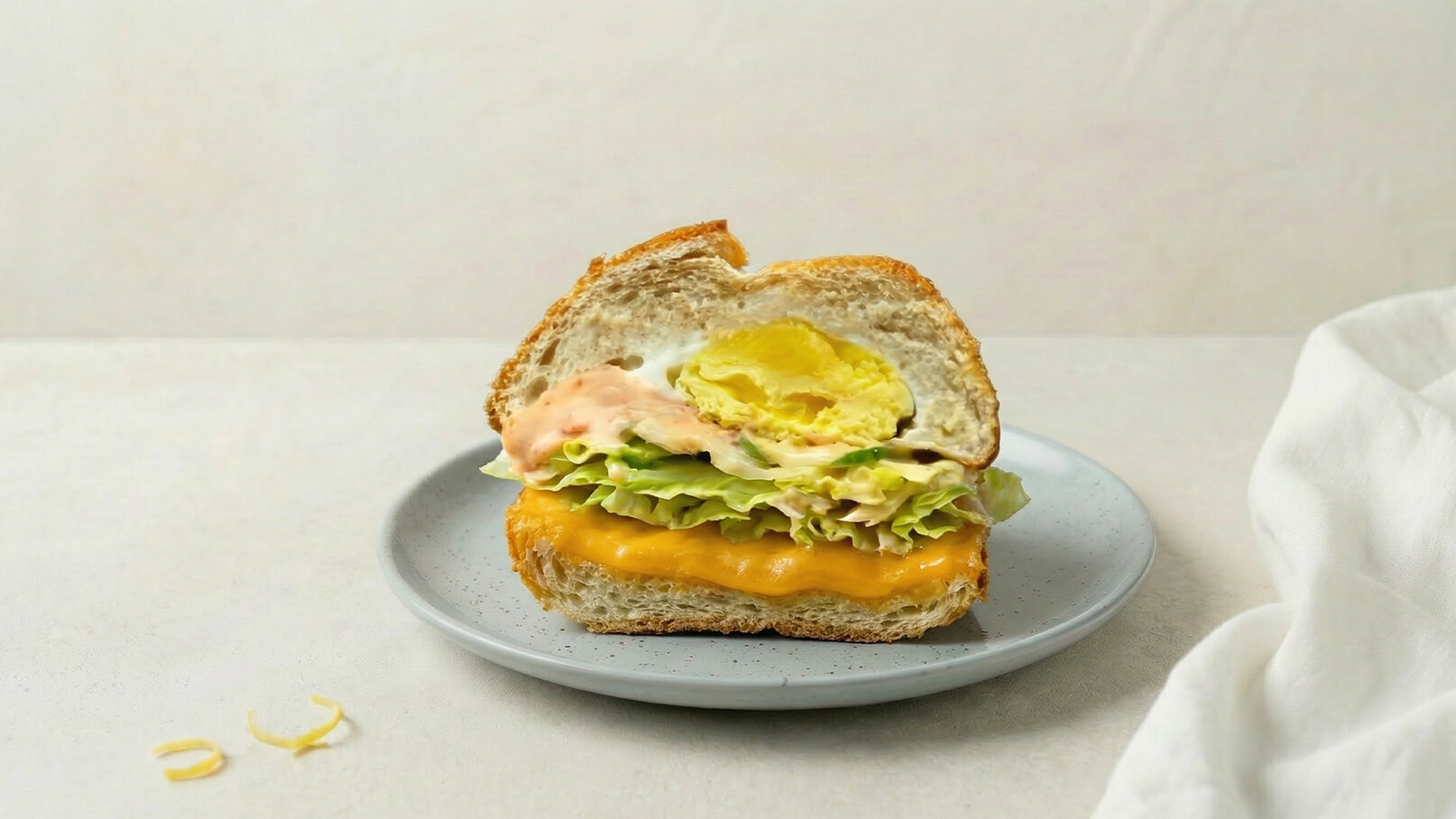 Ein halbierter Sandwich auf einem grauen Teller, gefüllt mit frischem Salat, einer Scheibe Käse und einer cremigen Sauce. Das Brötchen ist weich und leicht angeröstet, während die Füllung appetitlich aussieht. Der Hintergrund ist neutral und lenkt nicht von dem Sandwich ab.