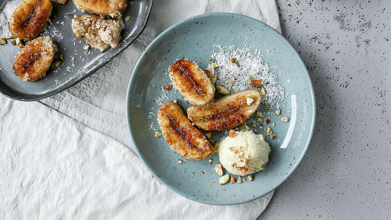 Ein hellblauer Teller mit gebratenen Bananen, die mit Kokosraspeln und gehackten Nüssen garniert sind. Neben den Bananen liegt eine Portion Vanilleeis. Im Hintergrund befindet sich eine dunkelgraue Servierplatte mit ähnlichen Bananen.