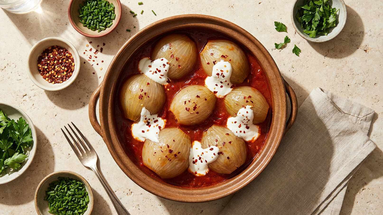 Eine Schüssel mit gefüllten Zwiebeln in Tomatensoße, garniert mit Sahne und roten Chiliflocken. Um die Schüssel herum befinden sich frische Kräuter und Gewürze, darunter Schnittlauch und Petersilie.