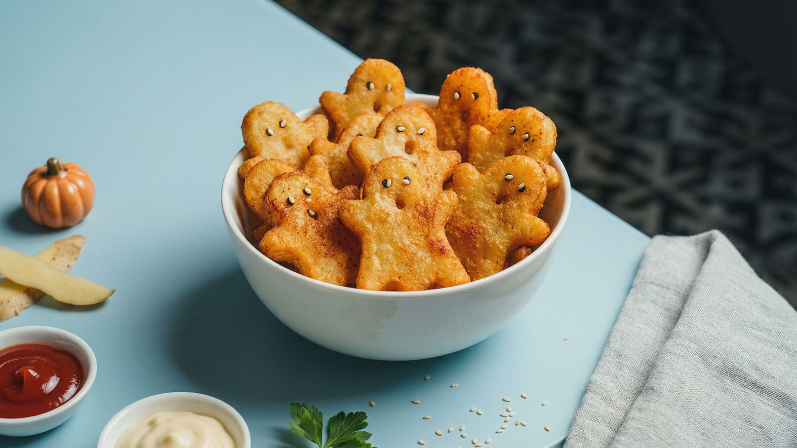 Eine weiße Schüssel voller knuspriger Snacks in Form von kleinen, gesichteten Männchen. Die Snacks sind goldbraun und haben schwarze Punkte als Augen. Neben der Schüssel liegen verschiedene Soßen in kleinen Schalen.