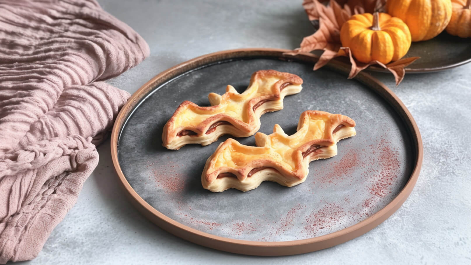Auf einem runden Teller liegen zwei gebackene Gebilde in Form von Fledermäusen. Die Teigstücke sind goldbraun und haben Füllungen. Im Hintergrund sind Deko-Elemente wie kleine Kürbisse und eine Stoffserviette zu sehen.