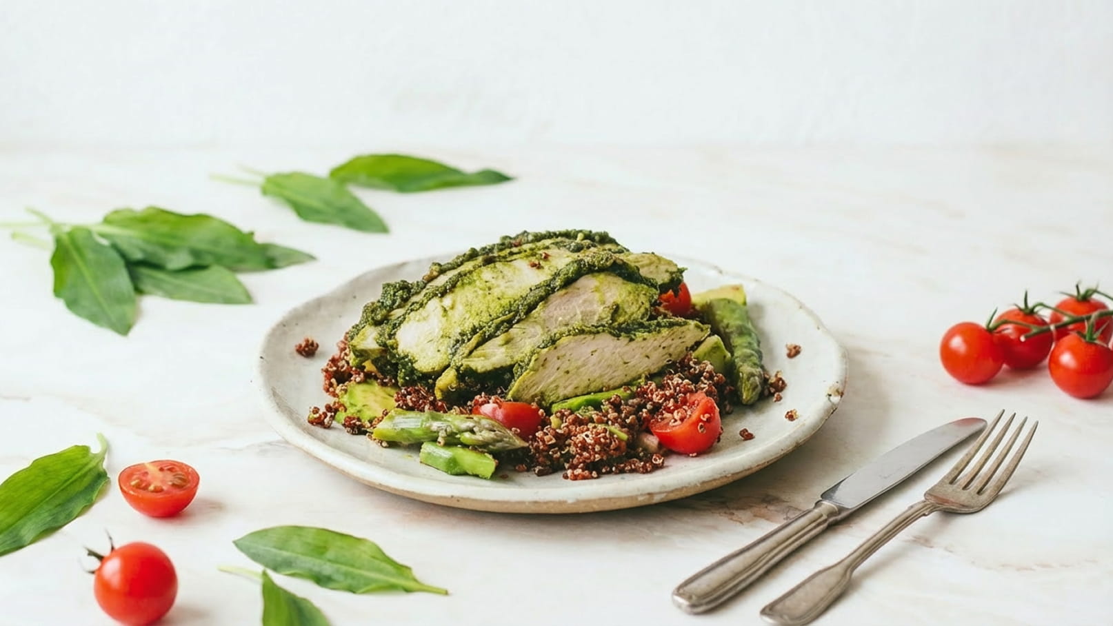 Ein Teller mit gegrilltem Hähnchen, umhüllt von grüner Kräuterpaste, serviert auf einem Bett aus Quinoa und frischen Gemüsesorten. Rote Kirschtomaten und grüne Blätter ergänzen die Präsentation. Das Gericht ist ansprechend angerichtet und vermittelt einen gesunden, frischen Eindruck.