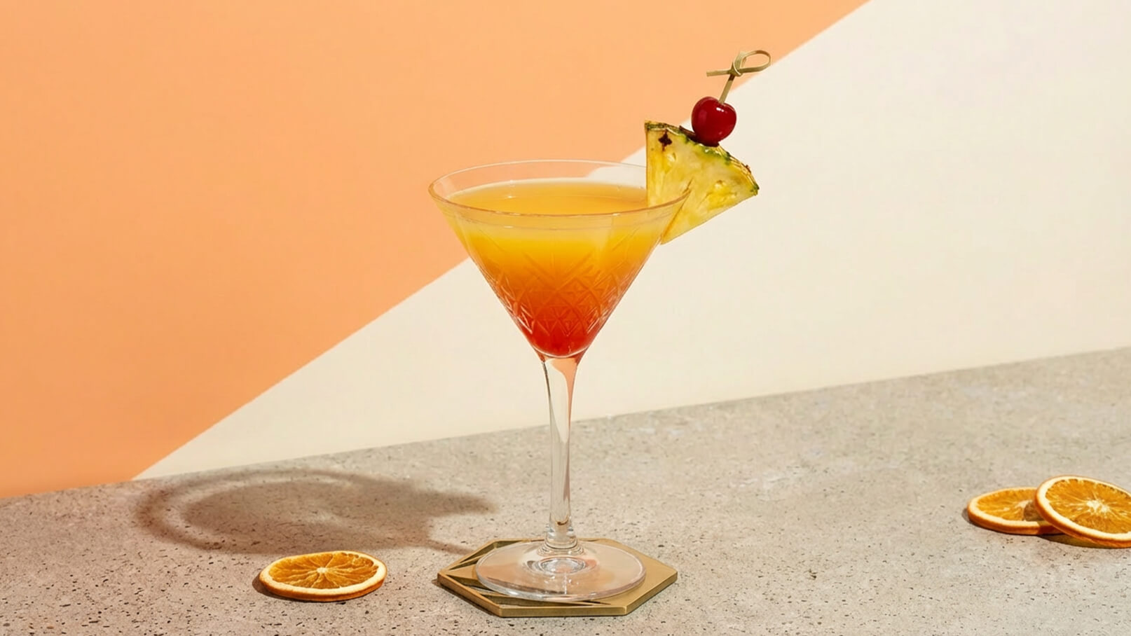 Ein farbenfroher Cocktail in einem eleganten Glas, kombiniert aus Gelb und Orange mit einem Stück Ananas und einer Kirsche als Garnitur. Im Hintergrund sieht man eine schlichte, helle Wand und einige Orangenscheiben auf der Tischoberfläche.