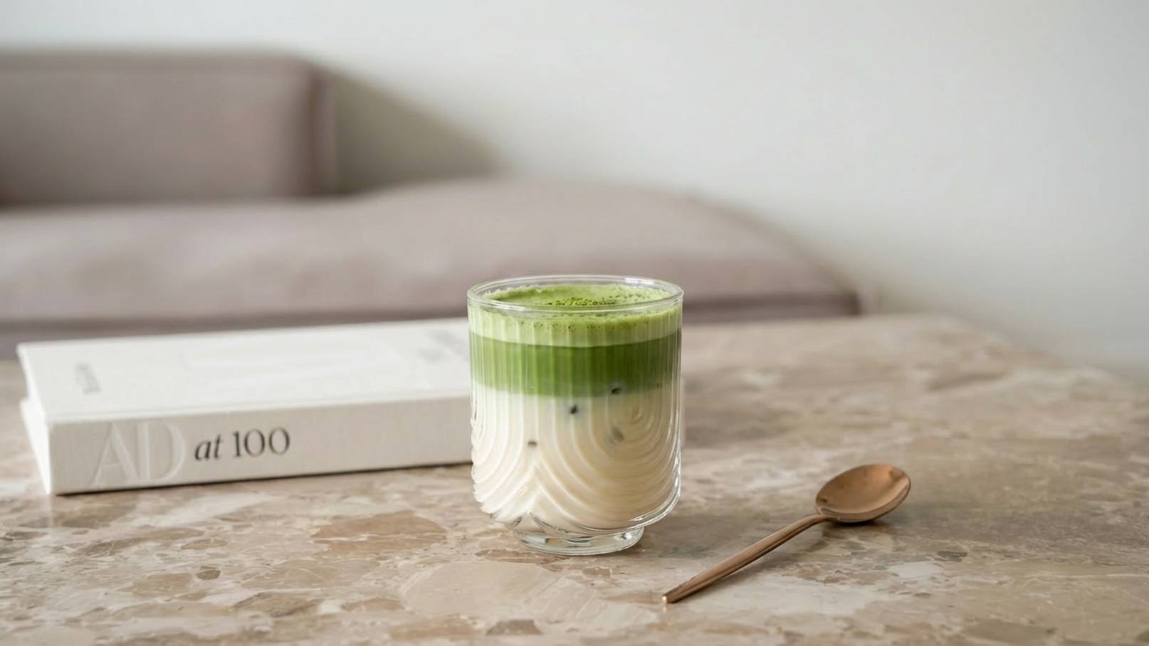 Ein transparentes Glas mit geschichtetem Getränk, bestehend aus einer hellen unteren Schicht und einer grünen oberen Schicht. Daneben liegt ein kleiner Löffel auf einem marmorierten Tisch, und im Hintergrund ist ein Buch aufgeschlagen.