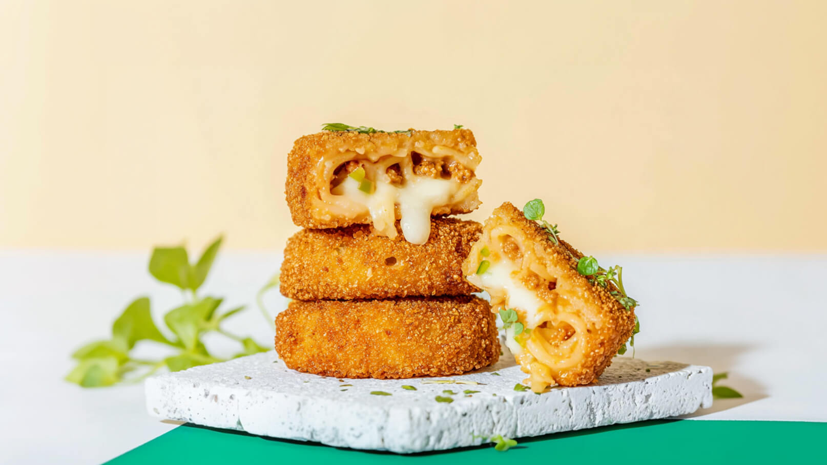Drei goldbraun frittierte Snacks sind übereinander gestapelt, von denen einer aufgeschnitten ist und geschmolzenen Käse sowie Pasta zeigt. Die Snacks liegen auf einem weißen Untergrund, umgeben von frischen Kräutern. Der Hintergrund ist in einem sanften Pastellton.