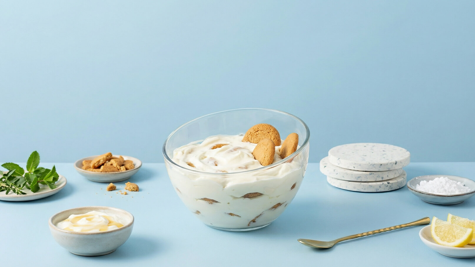 Eine Glasschüssel mit einer cremigen Nachspeise, garniert mit Keksen, steht im Mittelpunkt. Um die Schüssel herum liegen kleine Schalen mit Joghurt, Pfefferminzblättern und Zucker. Im Hintergrund sind Platten und Zitronenscheiben zu sehen. Der Hintergrund ist in sanftem Blau gehalten.