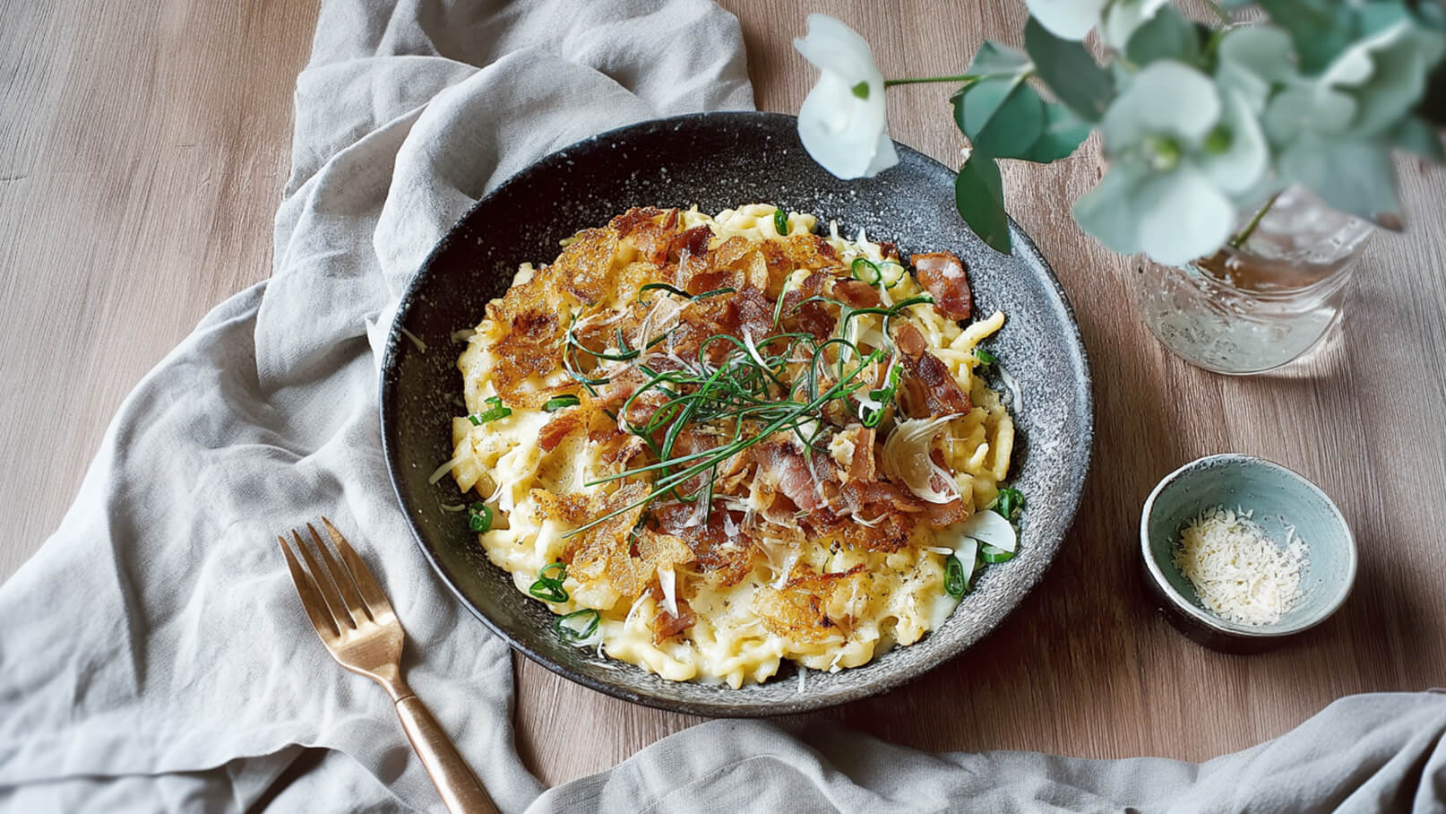 Eine dunkle Schüssel mit frisch zubereiteten Nudeln, die mit knusprigem Speck und frischen Kräutern garniert sind. Neben der Schüssel befindet sich ein kleiner Teller mit geriebenem Käse. Der Hintergrund zeigt eine Tischdecke und eine Vase mit Blumen.