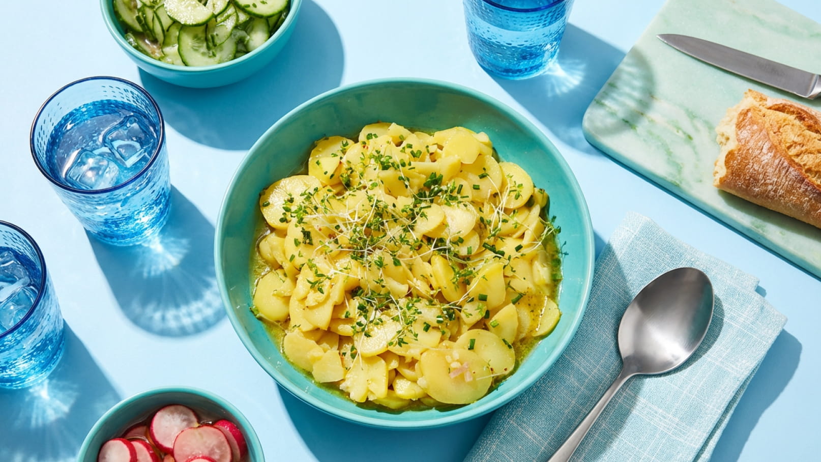 Eine grüne Schüssel mit warmem Kartoffelsalat, garniert mit frischen Kräutern, steht auf einem blauen Tisch. Daneben sind Gläser mit Wasser, eine kleine Schüssel mit Gurkenscheiben und eine andere mit Radieschen. Ein Stück Baguette liegt auf einem Teller.