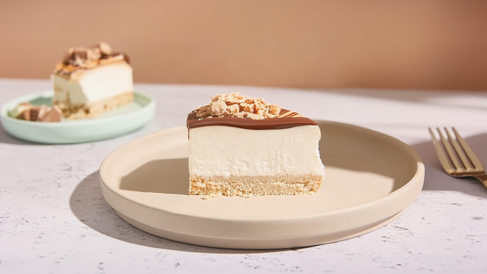Ein Stück Käsekuchen auf einem hellen Teller, mit einer glänzenden Schokoladenglasur und Knusperstückchen obendrauf. Im Hintergrund ein weiterer, ähnlicher Kuchen auf einem grünen Teller. Der Tisch hat eine helle, neutrale Oberfläche.