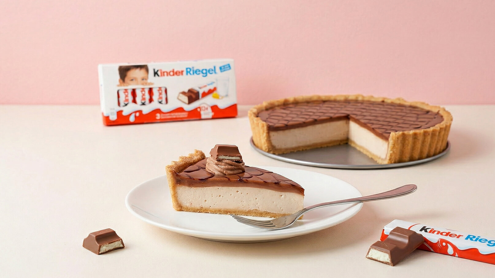 Ein Stück Kuchen mit einer cremigen Füllung und Schokoladendecke liegt auf einem weißen Teller mit einem Löffel daneben. Im Hintergrund steht eine ganze Torte, und eine Verpackung von Kinder Riegel ist sichtbar. Der Hintergrund ist in sanften Pastellfarben gehalten.