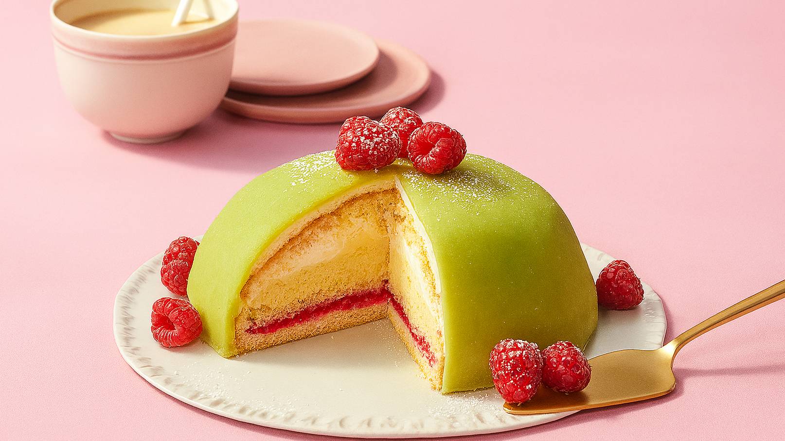 Ein rundes, geschnittenes Törtchen mit grünem Marzipanüberzug, gefüllt mit Schichten von Biskuit, Sahne und Himbeermarmelade. Oben sind frische Himbeeren dekoriert. Im Hintergrund steht eine Tasse Kaffee auf einem rosa Untergrund.