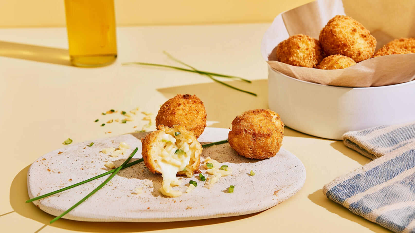 Auf einer hellen Fläche liegen mehrere goldbraune, panierte Bällchen. Eines ist aufgeschnitten und zeigt eine cremige Füllung. Im Hintergrund sind frische Schnittlauchhalme und eine Schale mit weiteren Bällchen zu sehen.