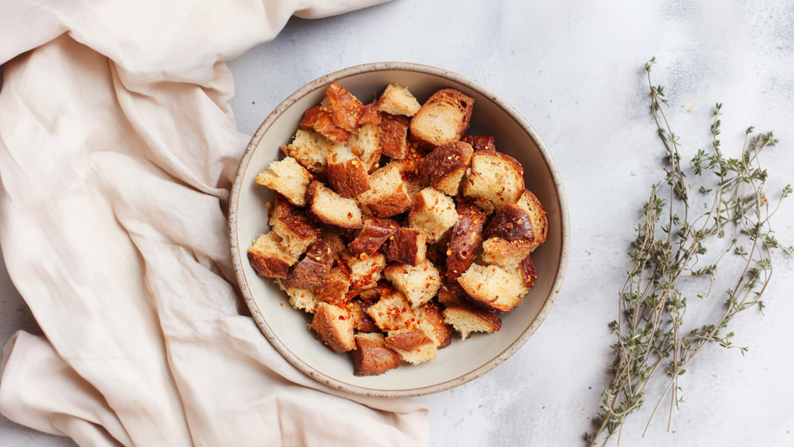Eine Schale mit goldbraun gerösteten Brotwürfeln ist im Vordergrund sichtbar. Daneben liegt ein Stück helles Tuch. Im Hintergrund sind einige Zweige Thymian platziert, die eine rustikale Atmosphäre schaffen.