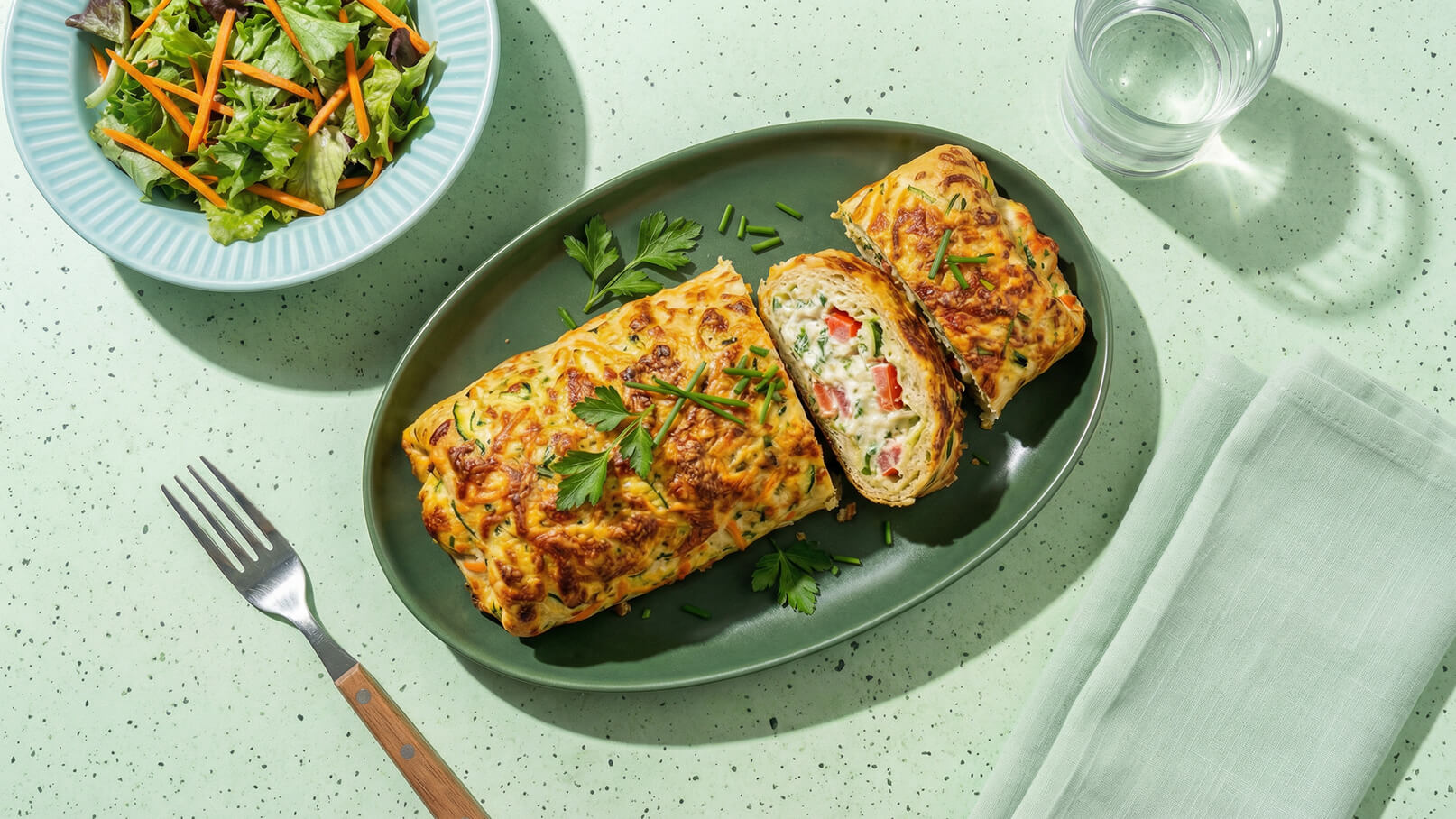 Auf einem grünen Teller liegt ein gefüllter Omelett-Roll, garniert mit Petersilie. Daneben befindet sich eine Schüssel mit gemischtem Salat und Möhrenstreifen sowie ein Glas Wasser. Ein Besteck liegt bereit.