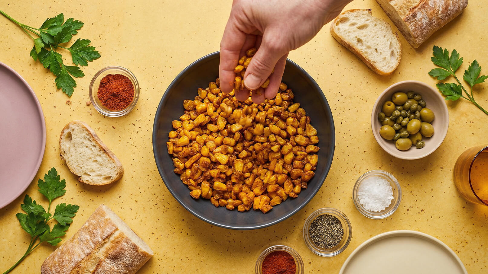Eine Hand streut Gewürze über eine Schüssel mit knusprig geröstetem Mais. Drumherum sind Zutaten wie Brot, Oliven, Gewürze und frische Kräuter auf einem hellen Tisch verteilt.