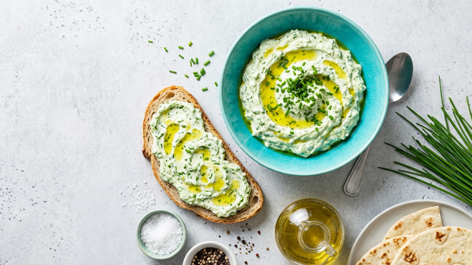 Ein blauer Teller mit cremiger Kräutercreme, garniert mit Schnittlauch und Olivenöl. Daneben liegt eine Scheibe Brot, die mit der Creme bestrichen ist. Im Hintergrund sind verschiedene Gewürze und frische Kräuter sichtbar.