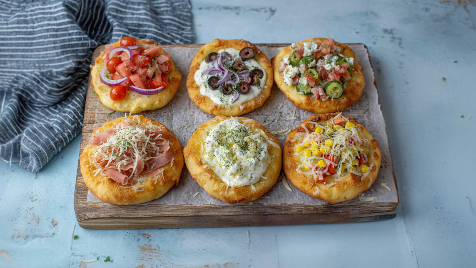 Sechs verschiedene belegte Fladenbrote auf einem Holzbrett. Die Beläge umfassen frische Tomaten, Oliven, Schinken, Käse, Gemüse und Mais. Die Fladenbrote sind knusprig und appetitlich angeordnet, mit Kräutern garniert. Ein graues Tuch liegt neben dem Brett.