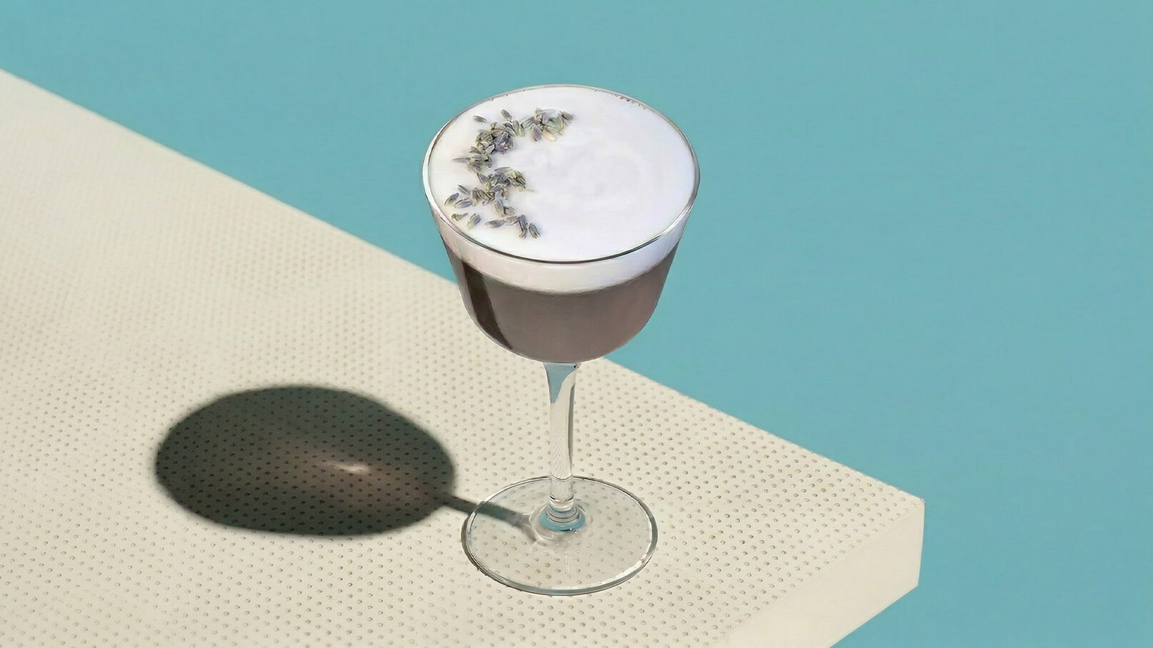 Ein elegantes Cocktailglas steht auf einer hellen, perforierten Oberfläche. Der Drink ist dunkel mit einer cremigen Schaumdecke, die mit kleinen Lavendelblüten dekoriert ist. Der Hintergrund ist in einem sanften Blau gehalten.