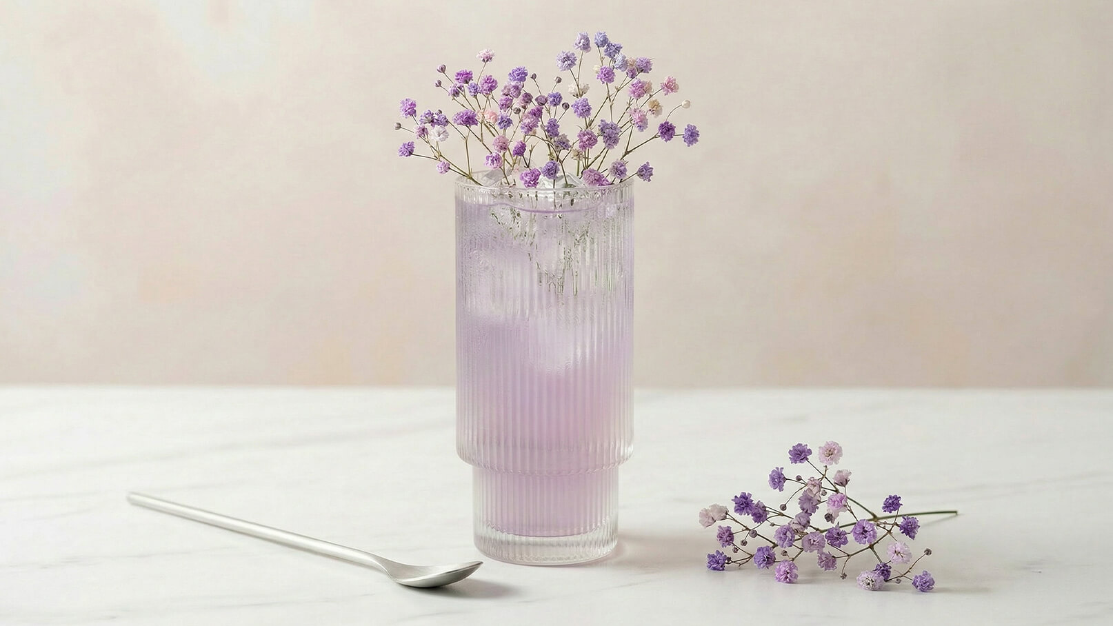 Ein durchsichtiger Becher mit einem lila Getränk, das Eiswürfel enthält, ist mit zarten lila Blumen dekoriert. Neben dem Becher liegt ein silberner Löffel auf einem weißen Tisch.