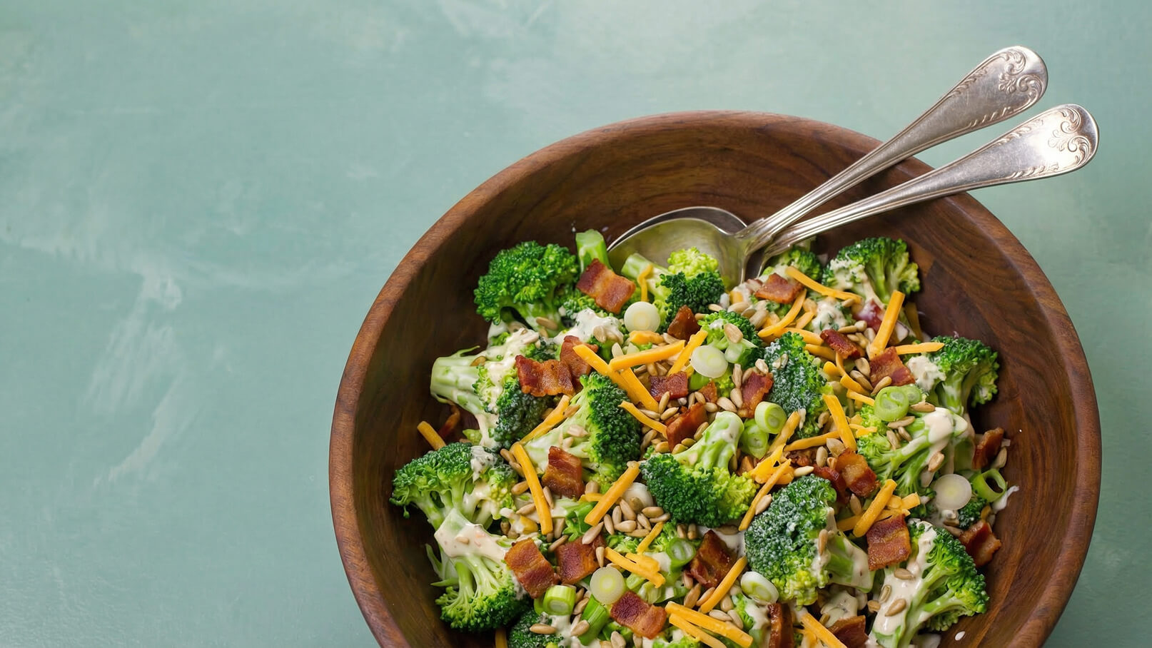 Eine hölzerne Schale enthält einen bunten Salat aus frischem Brokkoli, mit Speckstücken, Käse, Sonnenblumenkernen und Frühlingszwiebeln garniert. Zwei silberne Löffel liegen an der Seite der Schüssel. Der Hintergrund ist in sanften Grüntönen gehalten.