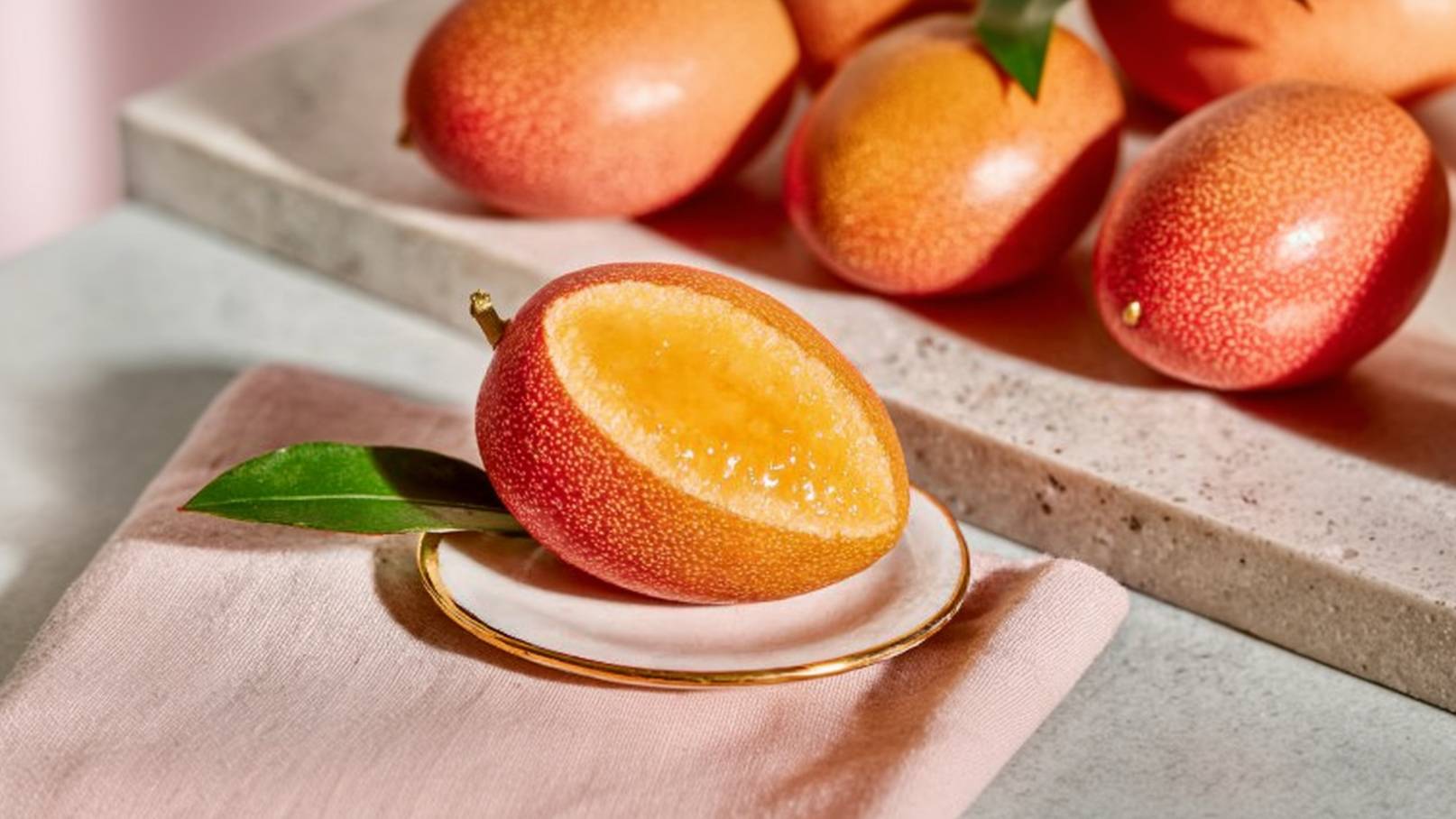 Ein auf einem Teller liegendes, halbiertes Obst, das leuchtend orangefarbenes Fruchtfleisch zeigt. Im Hintergrund sind mehrere ganze Früchte sichtbar. Das Obst hat eine glatte, rote Schale und liegt auf einem hellen Untergrund. Ein Grüner Blattansatz ist ebenfalls zu sehen.