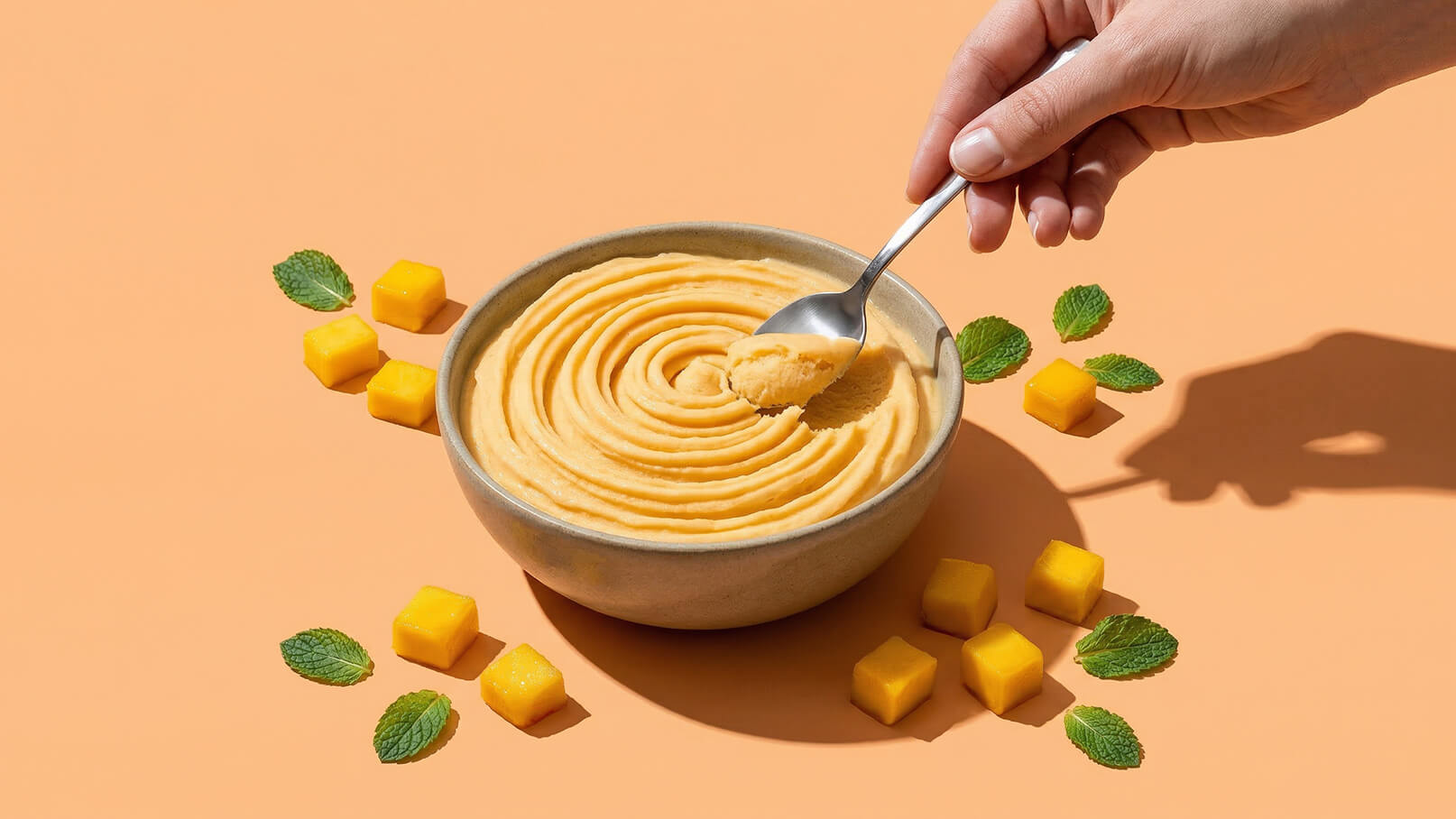Eine Hand hält einen Löffel und schöpft eine cremige, orangefarbene Mischung aus einer Schüssel. Der Teller ist von frischen Minzblättern und gelben Mango-Würfeln umgeben. Der Hintergrund ist ein einfarbiger, warmen Farbton.