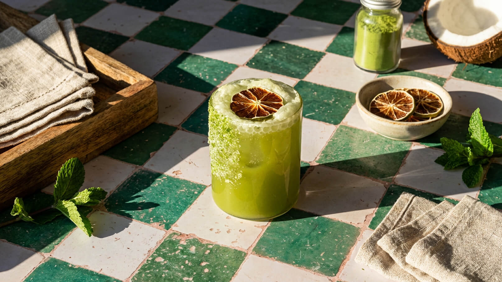 Ein Glas grüner Saft steht auf einem karierten Tisch. Der Saft ist mit Schaum und einer getrockneten Limette dekoriert. Neben dem Glas liegen frische Minzblätter und eine kleine Schale mit weiteren getrockneten Limetten. Im Hintergrund ist ein Holzbrett mit Servietten sichtbar.