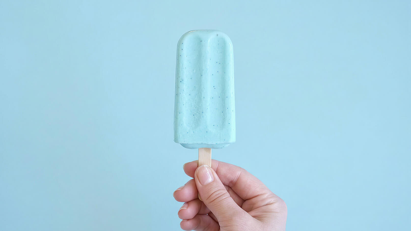 Eine Hand hält ein grünes Eis am Stiel mit einer glatten Oberfläche. Der Hintergrund ist einfarbig hellblau.