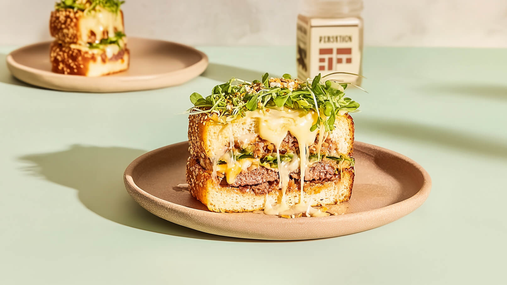 Ein saftiger Burger liegt auf einem Teller, mit geschmolzenem Käse, frischem Salat und Sprossen. Der Burger hat eine goldene Sesamkruste und eine dicke Fleischschicht, die aus dem bunten Gemüse hervorschaut. Im Hintergrund ist ein weiterer Burger auf einem anderen Teller zu sehen.