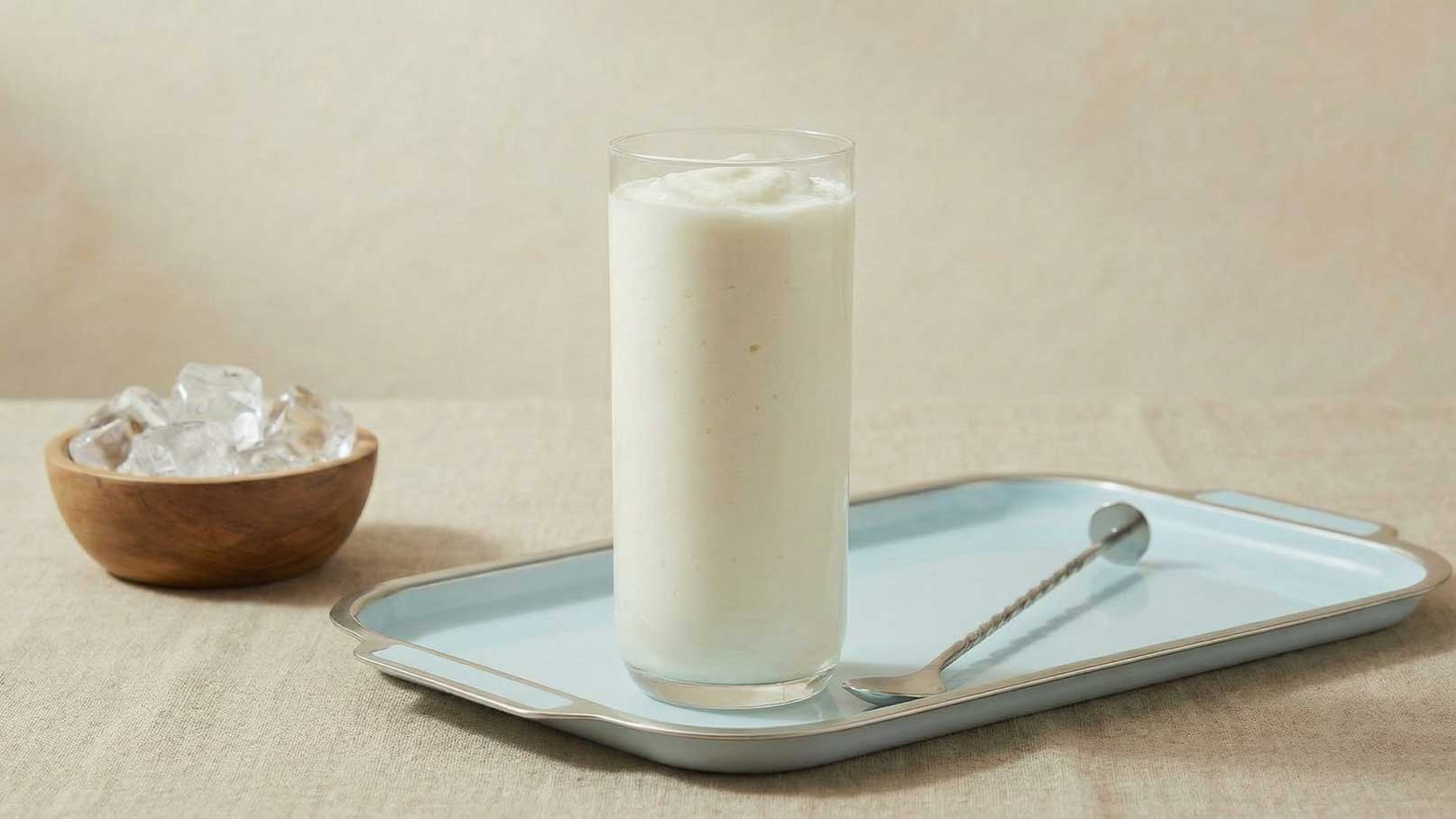 Ein Glas mit einem cremigen, weißen Getränk steht auf einem blauen Tablett. Neben dem Glas liegt ein Löffel. Im Hintergrund befindet sich eine Schale mit Eiswürfeln. Die Szene vermittelt eine erfrischende, einladende Atmosphäre.