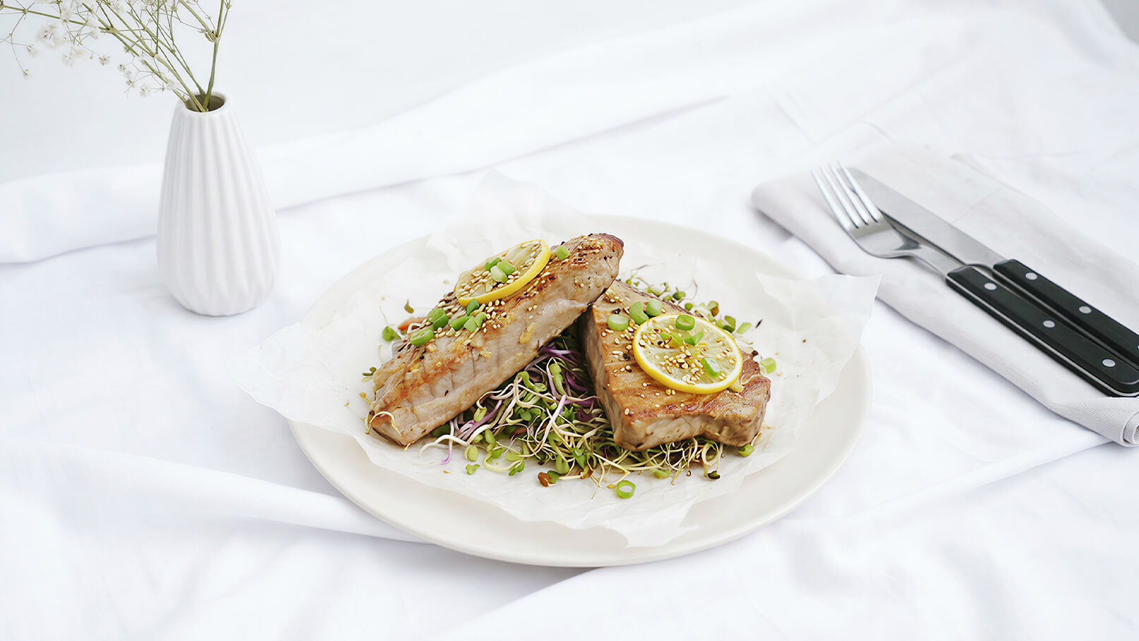 Auf dem Bild ist ein Teller mit zwei gegrillten Fischfilets zu sehen, die mit Zitronenscheiben belegt sind. Sie liegen auf einem Bett aus Sprossen. Im Hintergrund steht eine schlichte Vase mit Blumen auf einer weißen Tischdecke.
