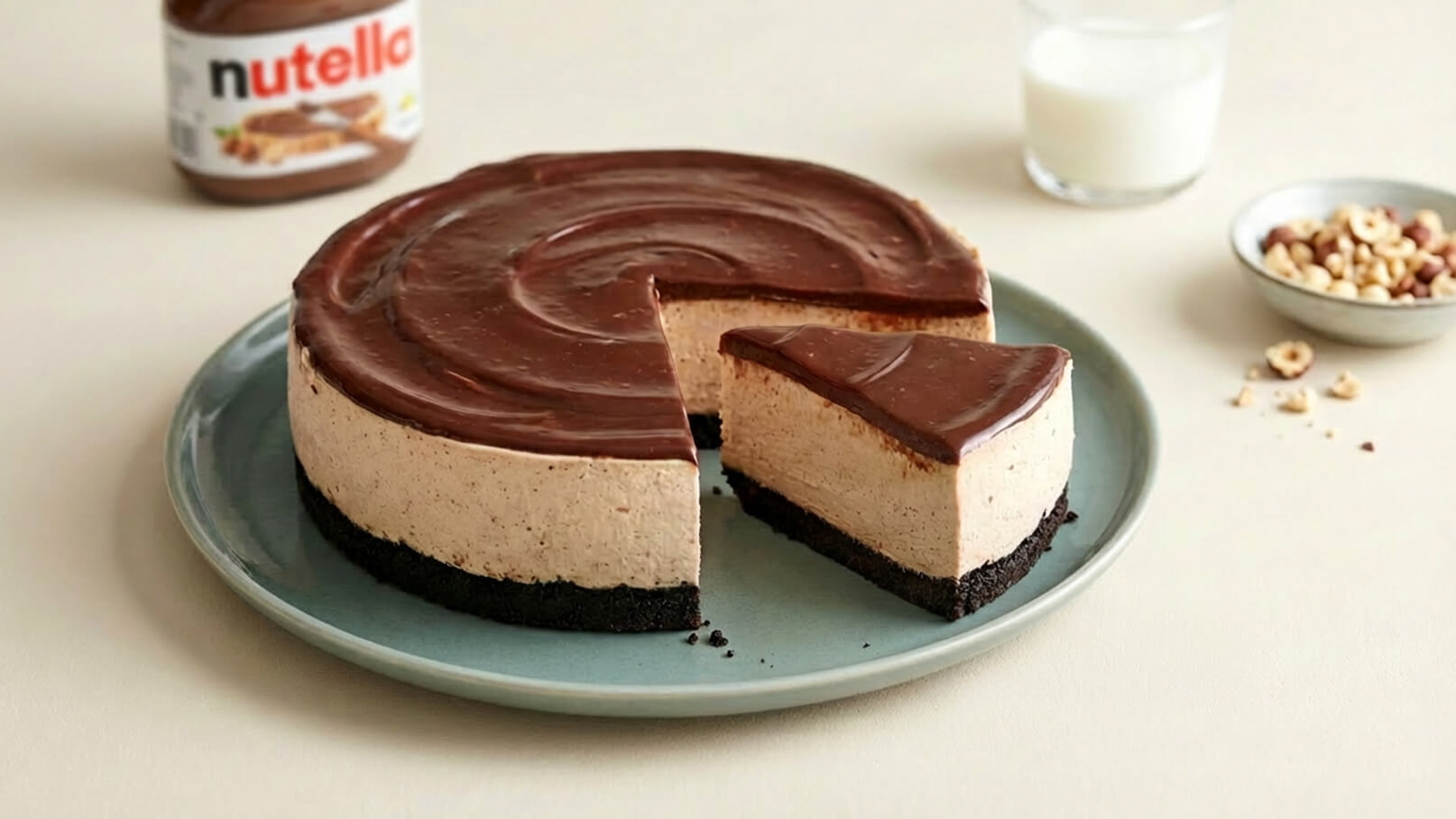 Ein praller Nutella-Käsekuchen auf einem blauen Teller. Ein Stück wurde herausgeschnitten, sodass die schichtartige Füllung und der schokoladige Belag sichtbar sind. Im Hintergrund befindet sich ein Glas Milch und eine kleine Schüssel mit Haselnüssen.