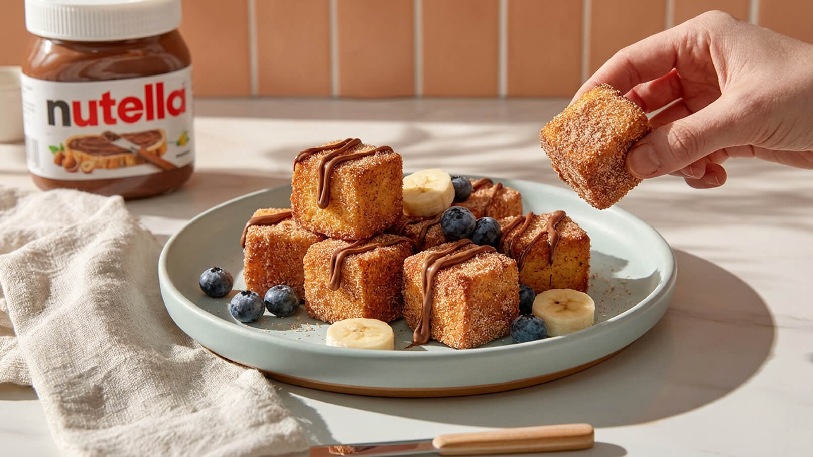 Ein Teller mit kleinen, goldbraunen Würfeln, die mit Zimt und Zucker bestreut sind, steht im Vordergrund. Einige sind mit Nutella verziert. Daneben liegen Blaubeeren und Bananenscheiben. In einer Hand wird ein Würfel zum Essen angehoben. Im Hintergrund steht ein Glas Nutella.