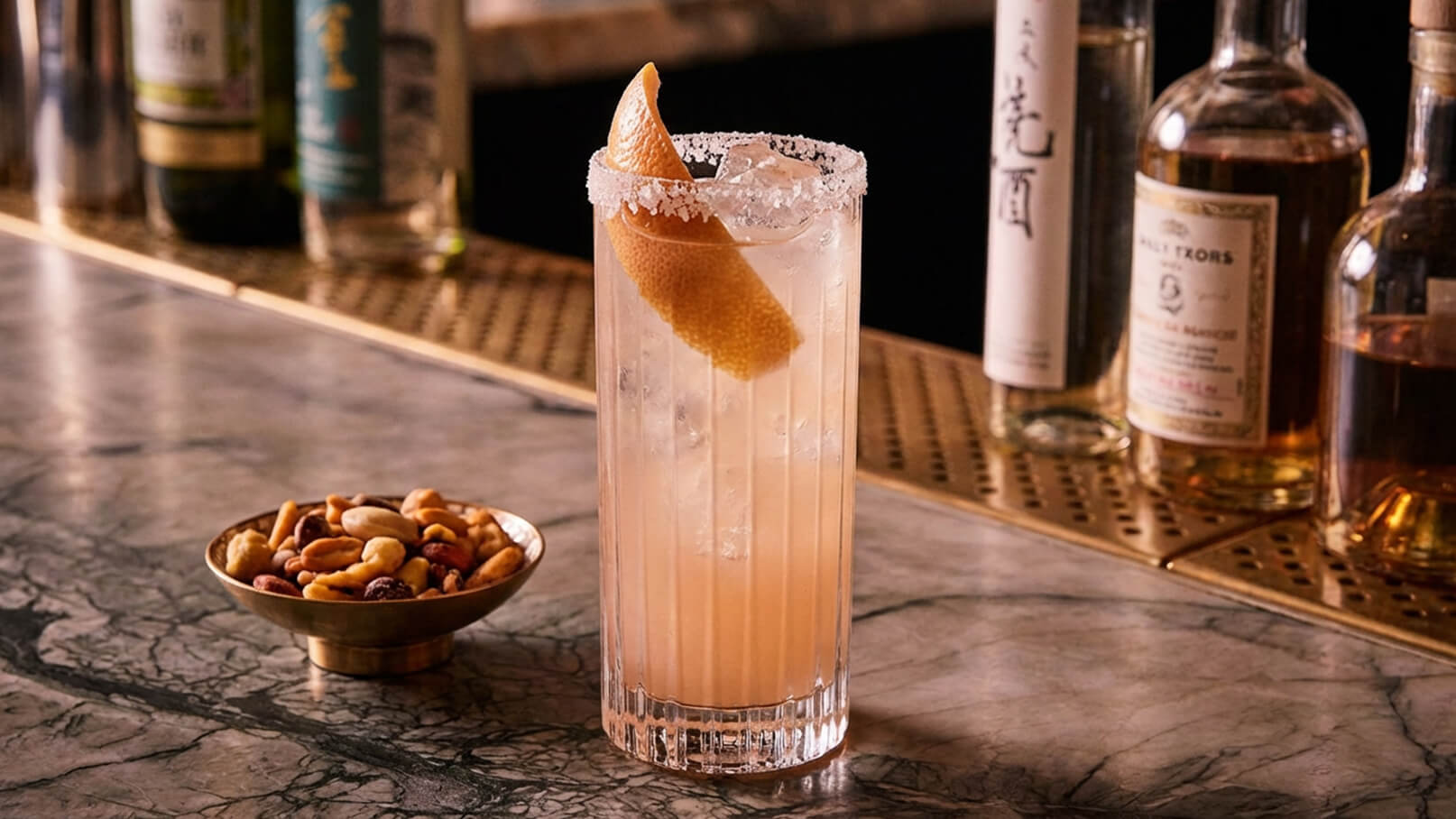 Ein hoher Glasbecher mit einem erfrischenden Cocktail steht auf einer Theke. Der Drink hat eine salzige Randverzierung und ist mit einer Orangenscheibe garniert. Neben dem Glas befindet sich eine kleine Schale mit gemischten Nüssen.