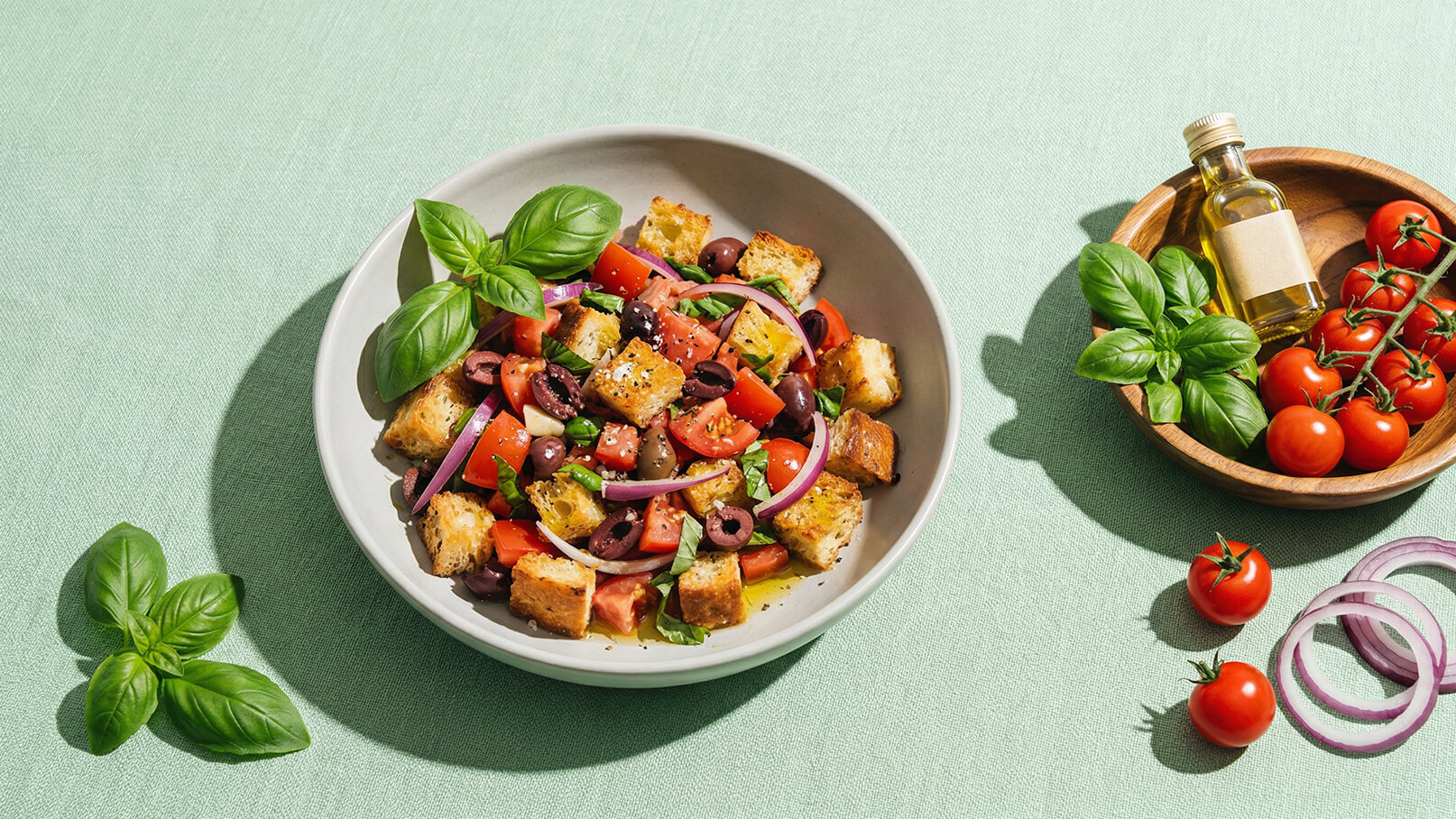 Eine Schüssel mit einem bunten Salat, bestehend aus gewürfeltem Brot, Tomaten, Gurken, roten Zwiebeln und Oliven, garniert mit frischem Basilikum. Daneben befinden sich eine Schale mit Kirschtomaten und eine Flasche Olivenöl auf einem hellen Untergrund.