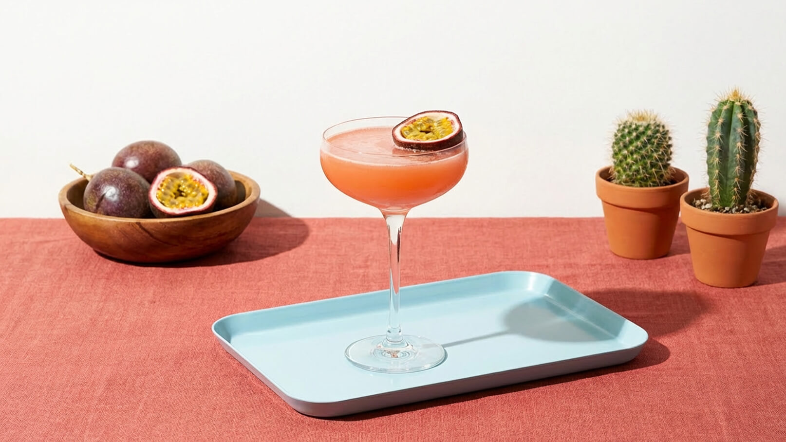 Ein elegantes Cocktailglas mit pinkfarbener Flüssigkeit und einer Passionsfrucht-Scheibe steht auf einem blauen Tablett. Im Hintergrund befindet sich eine Schale mit Passionsfrüchten und zwei kleine Töpfe mit Kakteen auf einem roten Tisch.