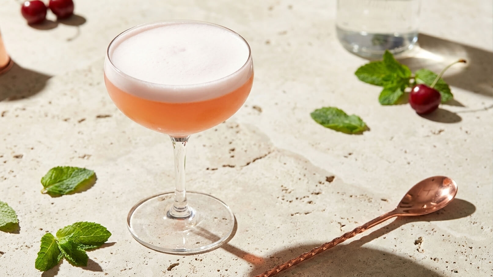 Ein elegantes Glas mit pinkfarbenem Cocktail, das eine cremige Schaumkrone hat, steht auf einem hellen Steinuntergrund. Um das Glas liegen frische Minzblätter und ein paar Kirschen, während ein Kupferlöffel daneben platziert ist.