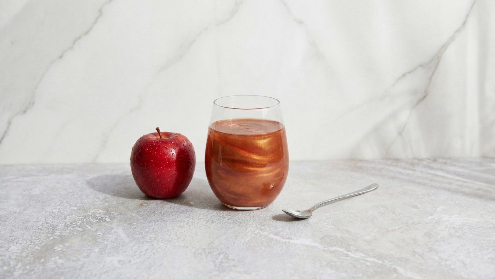Ein roter Apfel liegt neben einem Glas mit einer braunen, schimmernden Flüssigkeit. Ein kleiner Löffel befindet sich neben dem Glas. Der Hintergrund ist hell und marmoriert.