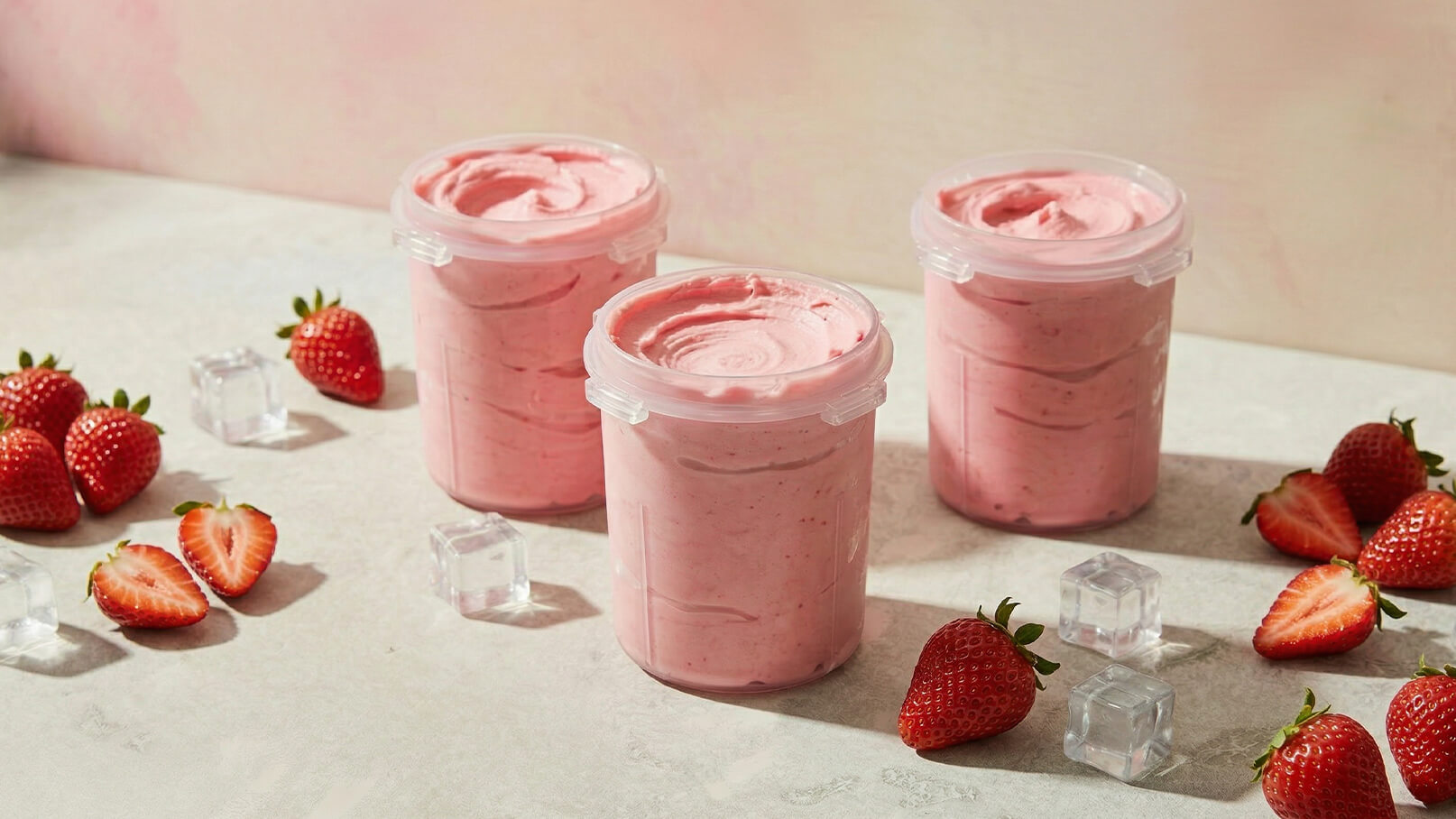 Drei transparente Behälter mit rosa Erdbeereis stehen auf einer Tischoberfläche. Um die Behälter herum liegen frische Erdbeeren und einige Eiswürfel. Der Hintergrund ist sanft gefärbt und verleiht eine appetitliche Atmosphäre.
