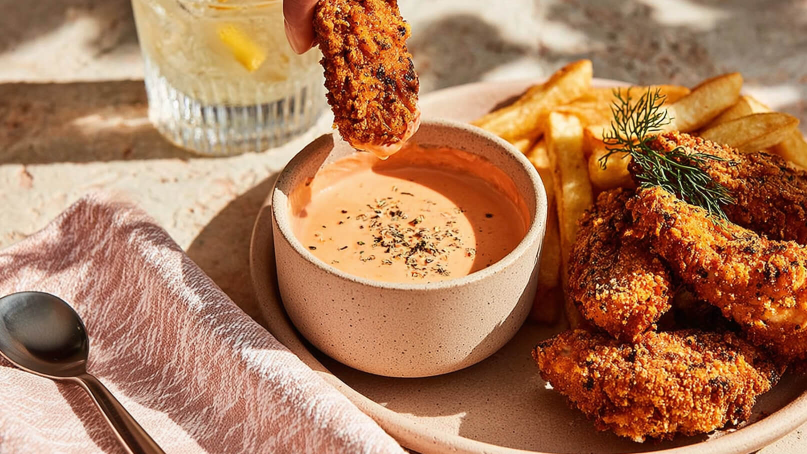 Ein Teller mit knusprigen Hähnchenstücken liegt neben einer kleinen Schüssel mit roter Sauce, in die ein Hähnchenstück getaucht wird. Pommes frites sind auf der Seite angeordnet. Der Tisch ist hell und leicht strukturiert.