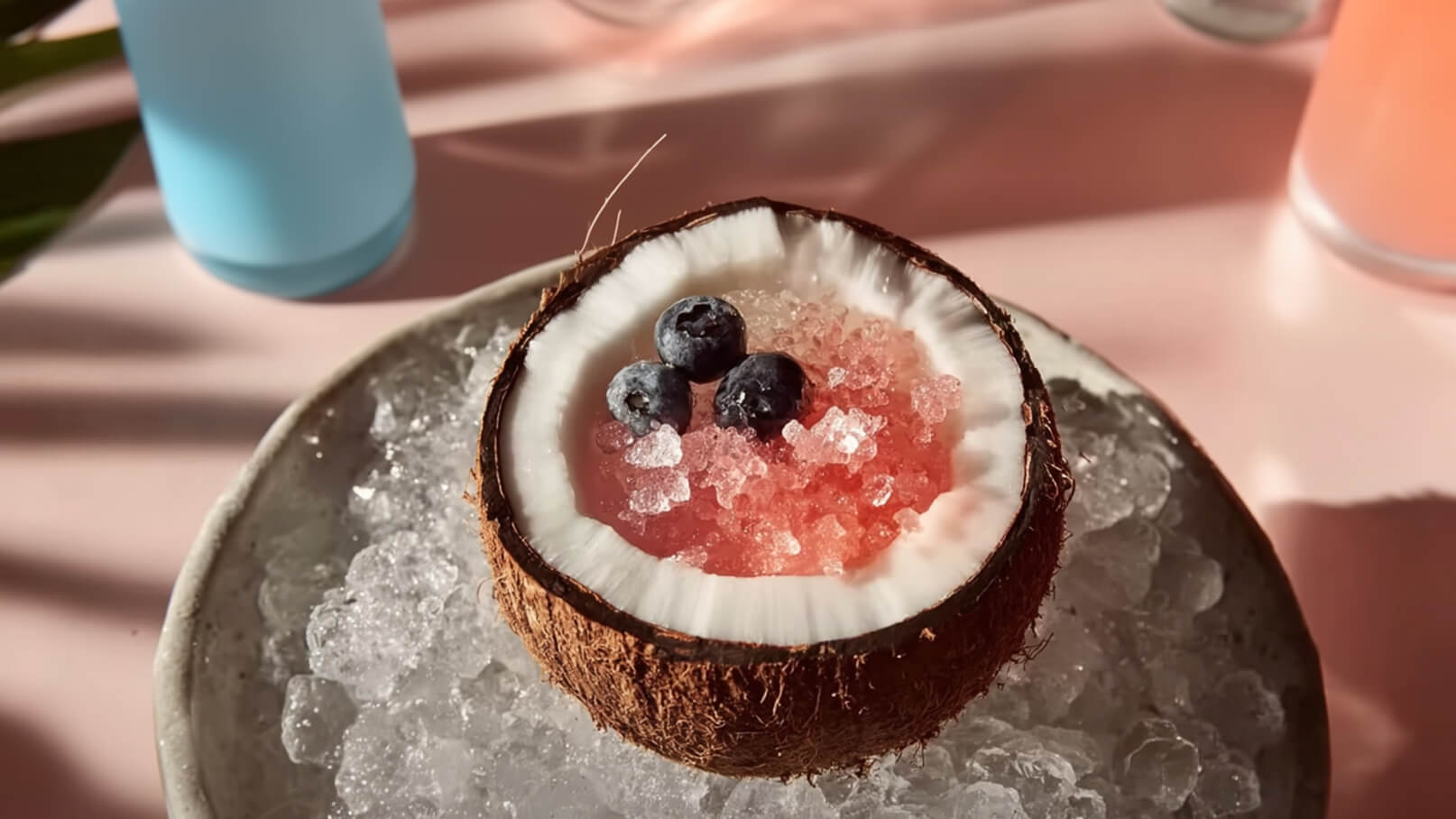 Eine halbe Kokosnuss ist auf einem Teller mit Eis platziert. In der Kokosnuss befinden sich gefrorene rote Stücke und drei Heidelbeeren. Der Hintergrund ist leicht verschwommen und zeigt eine entspannte Atmosphäre.
