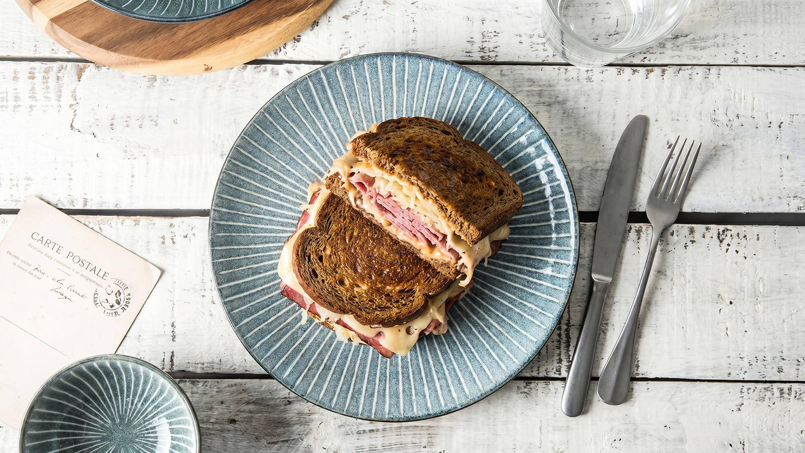Auf einem blauen Teller liegt ein Sandwich mit mehreren Schichten. Die Brotscheiben sind dunkel und knusprig, gefüllt mit Aufschnitt und Käse. Daneben liegen Besteck und ein weiteres Geschirrteil auf einem rustikalen Holztisch.