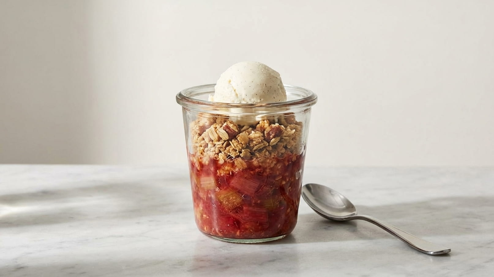 Ein Glas mit Rhabarber-Crumble, überzogen mit einer Kugel Vanilleeis. Der Crumble ist leicht goldbraun, und die fruchtige Füllung ist sichtbar. Neben dem Glas liegt ein Löffel auf einer Marmoroberfläche.