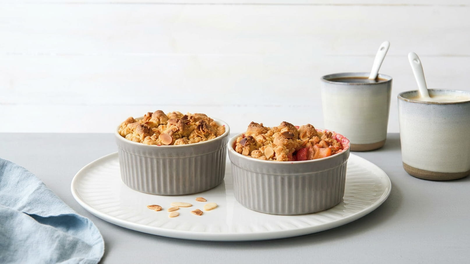 Zwei grau gestreifte Portionsschalen mit fruchtigem Crisp, überbacken mit einer knusprigen Haferflockenkruste, stehen auf einem weißen Teller. Im Hintergrund sind zwei helle Tassen sichtbar.