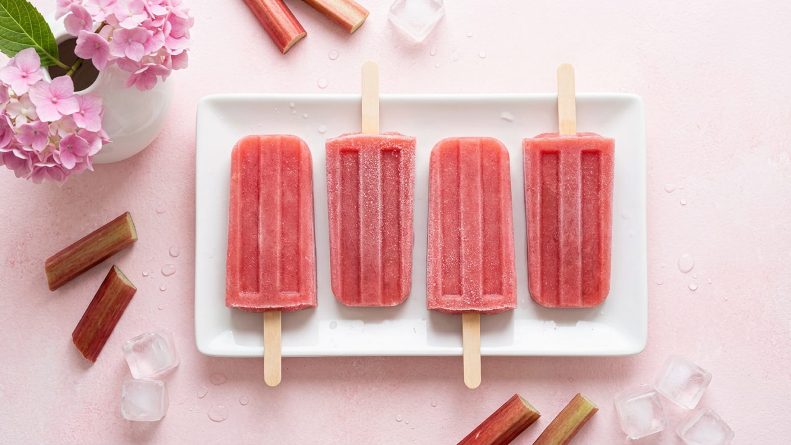 Auf einem weißen Teller liegen vier gefrorene Frucht-Eis am Stiel in kräftigem Rosaton. Um den Teller herum befinden sich Eiswürfel und einige Stücke Rhabarber. Im Hintergrund sind zartrosa Blüten zu sehen.